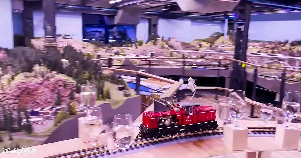 A red model train sits on tracks amidst a miniature landscape and dining setup.