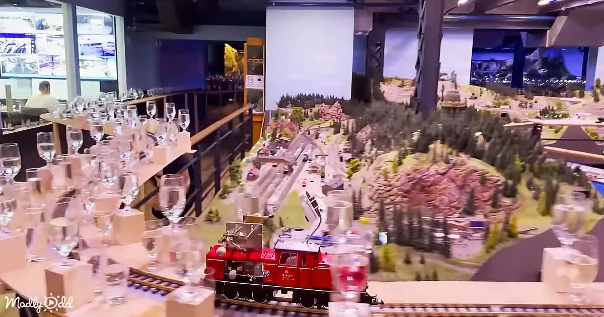 A red model train sits on tracks amidst a miniature landscape with trees and buildings, surrounded by wine glasses.