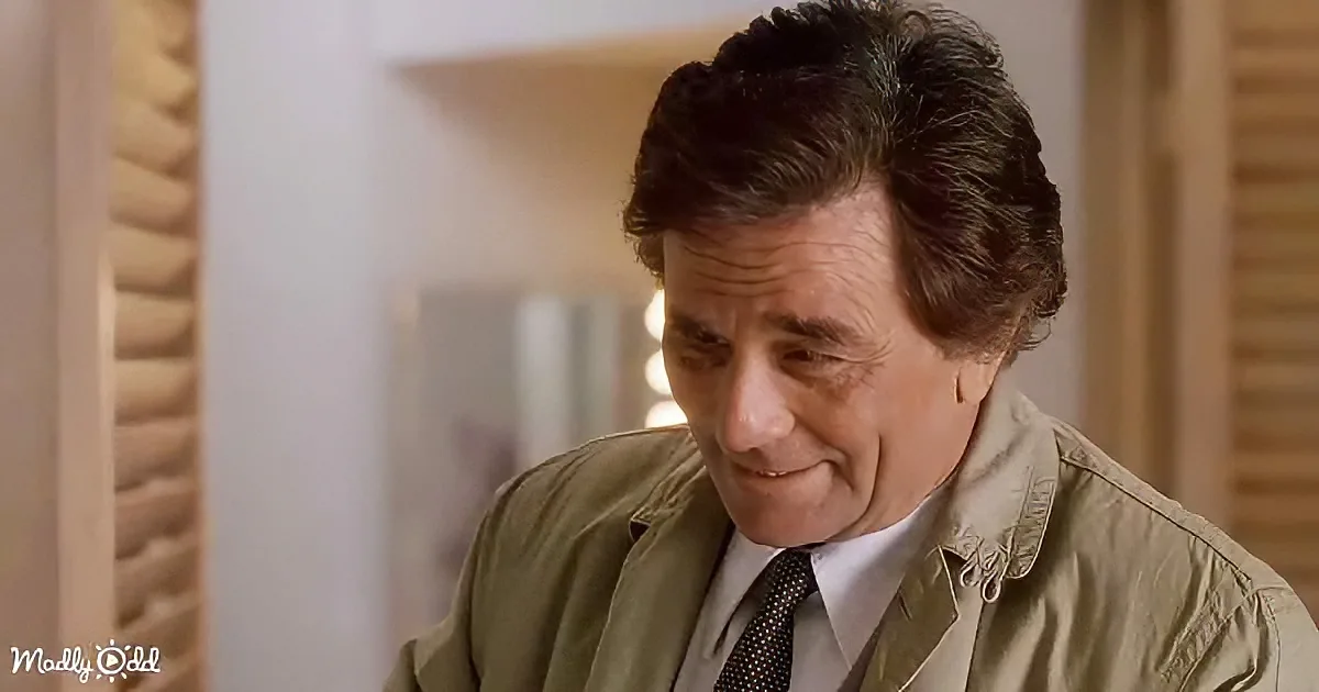 A man in a beige jacket and tie smiles while looking down, with a blurred background.