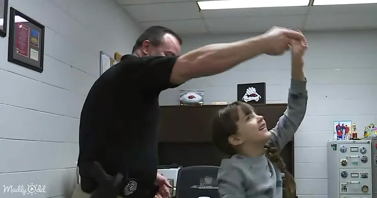 A man in a black shirt holds a young girl's arm up, both smiling in a room with white walls and trophies.