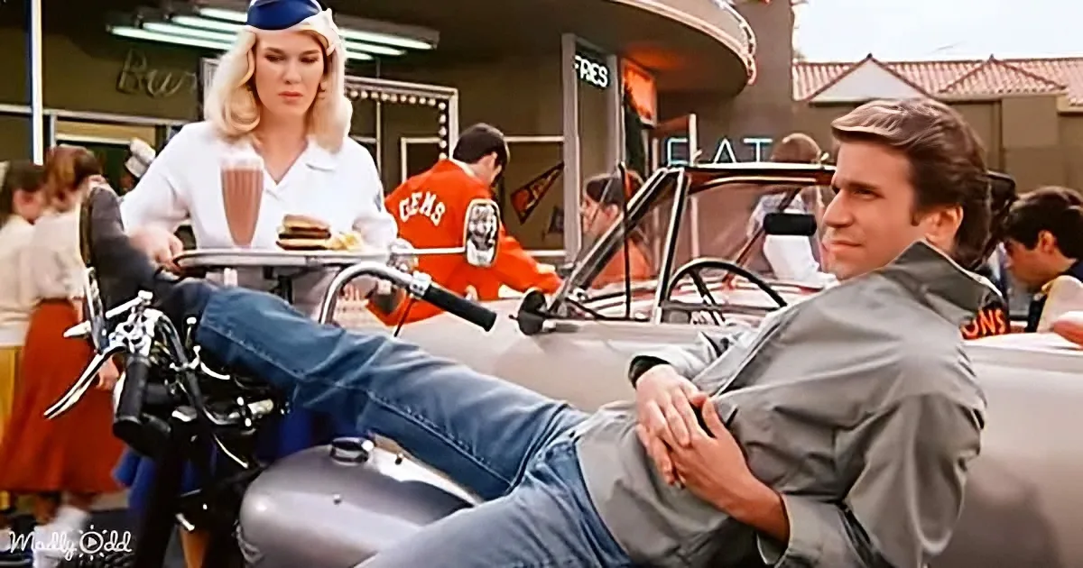 A man in a grey jacket and jeans sits on a motorcycle, holding a tray with a waitress in a diner.