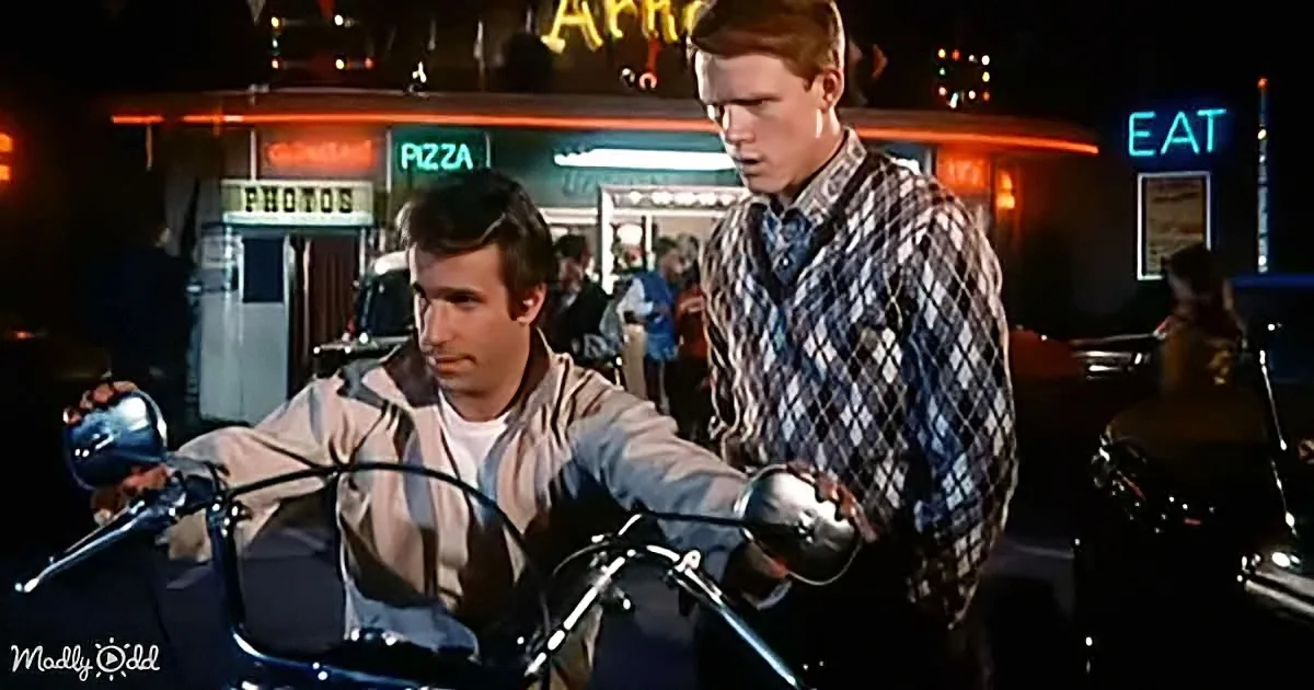 A man on a motorcycle interacts with another man in front of a neon-lit diner.