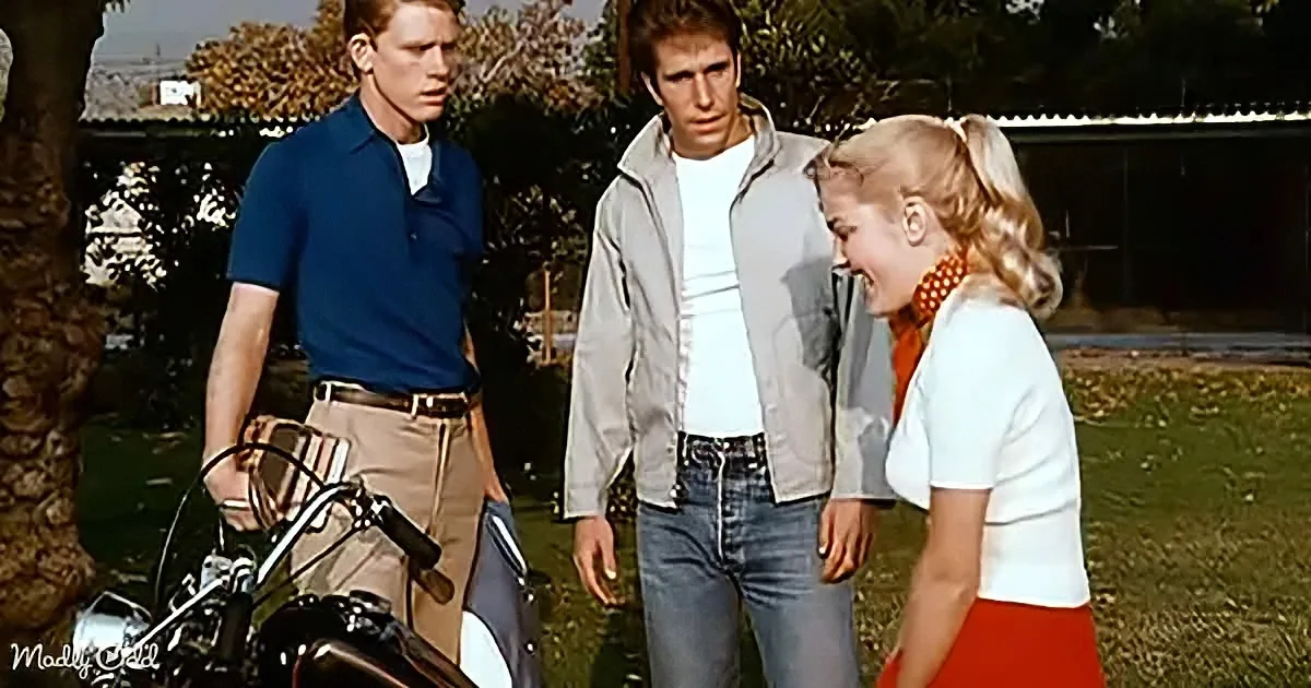 A man in a white shirt and gray jacket stands with two others, one in a blue shirt and khaki pants, another in a white...