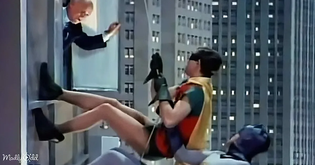 A woman in a superhero costume is hanging from a building, while another person reaches up to help her.