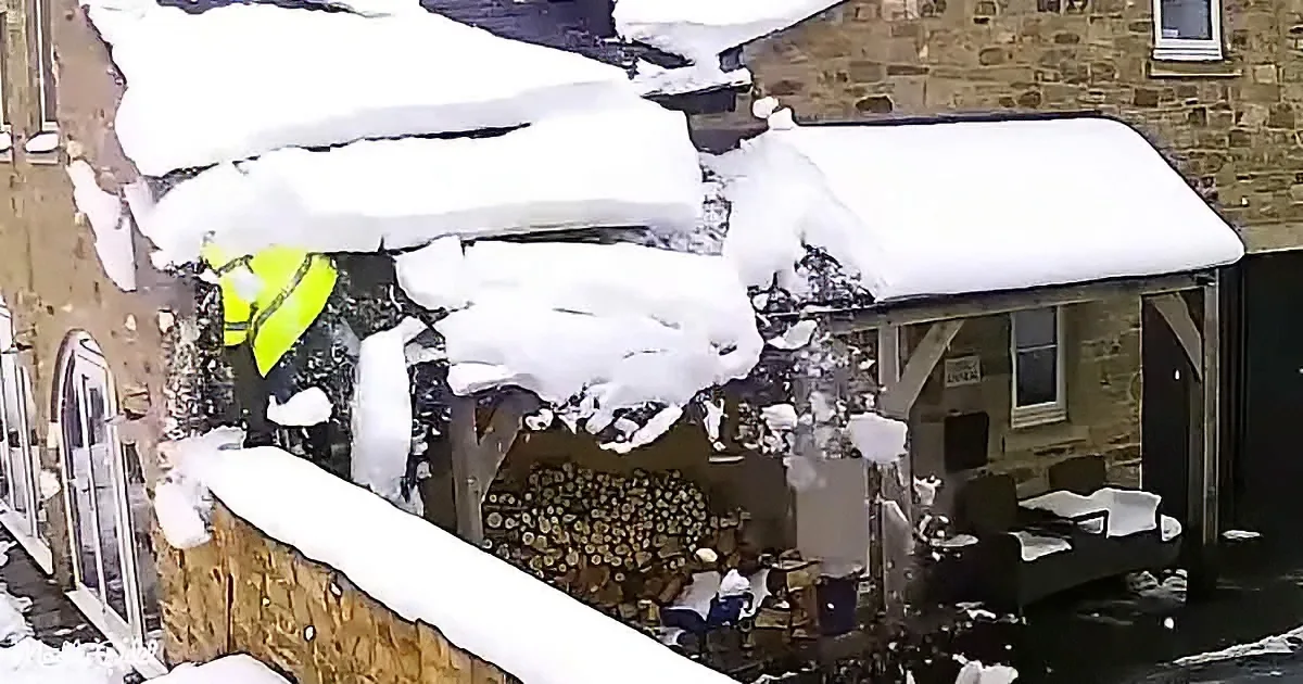 A person in a yellow jacket stands amidst a snowy scene with a large pile of wood and snow-covered buildings.