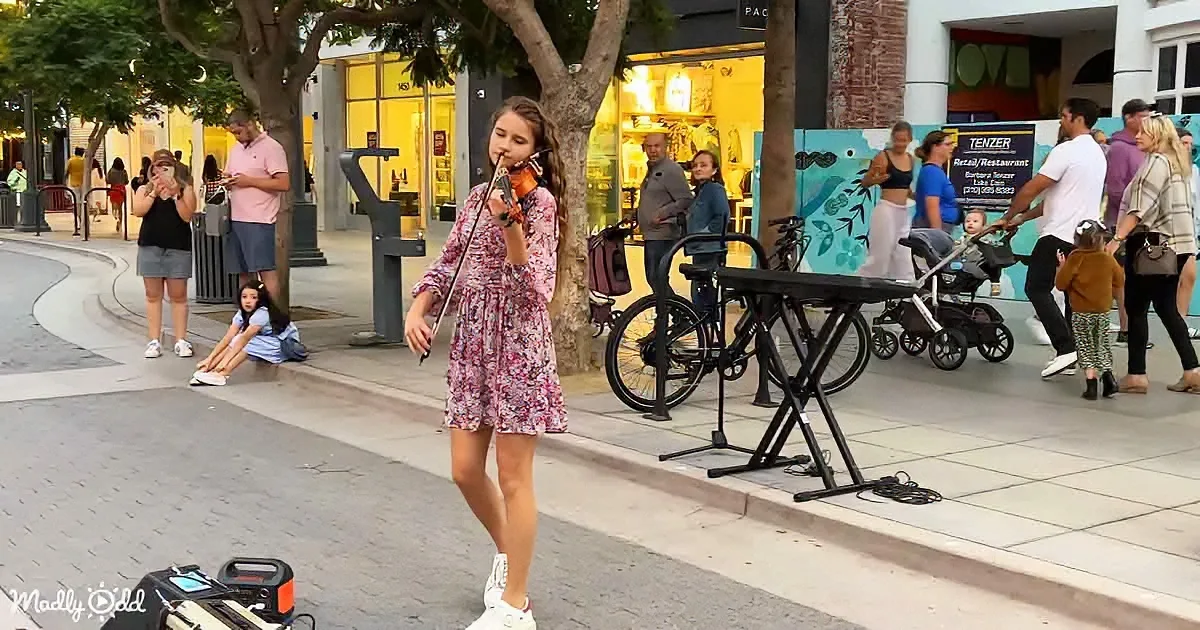 A girl in a floral dress plays violin on a sidewalk, surrounded by people and shops.