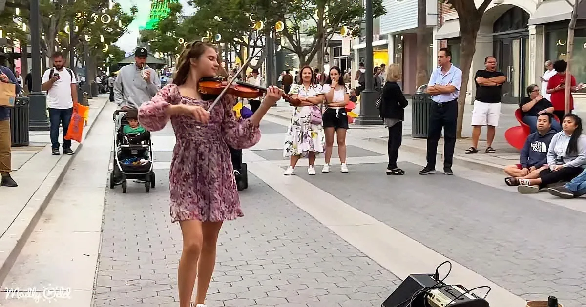 A woman in a floral dress plays the violin on a busy city sidewalk, surrounded by onlookers and passersby.