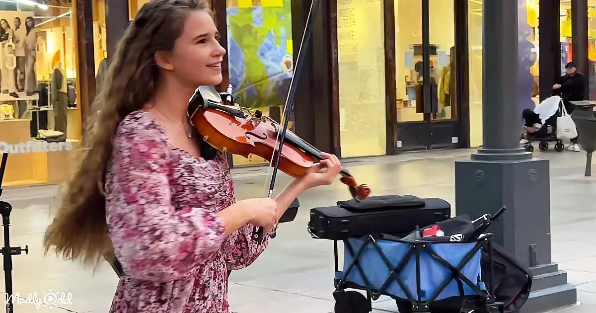 A woman in a floral dress plays the violin on a city street, smiling, with a case and stand nearby.