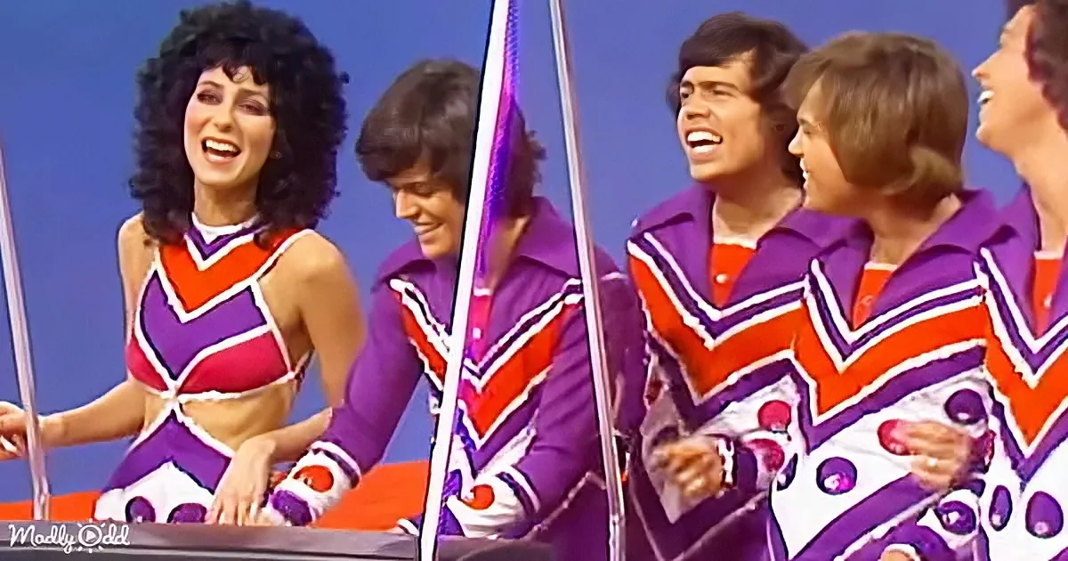 A woman in a colorful, retro outfit is smiling and posing with three men in matching purple and orange outfits.