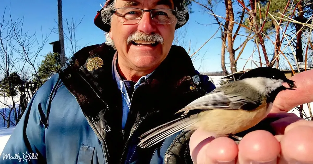 A man in a winter jacket holds a small bird on his hand, smiling in a snowy outdoor setting.