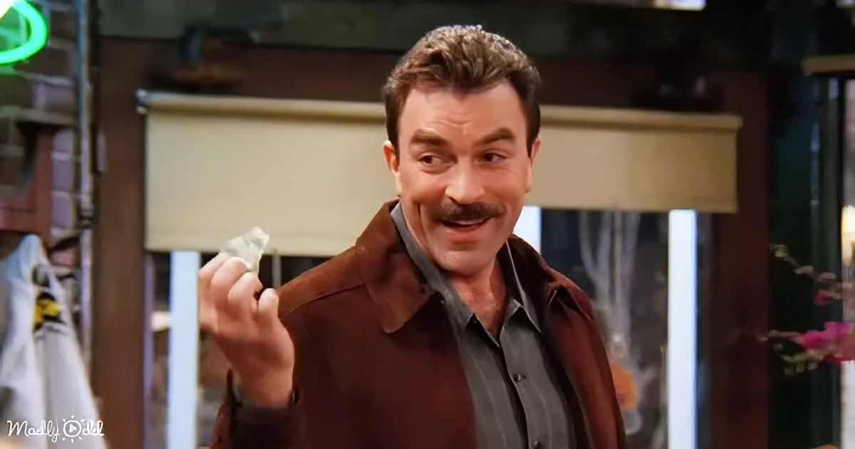 A man with a mustache and brown jacket holds a dollar bill, smiling in a dimly lit room.