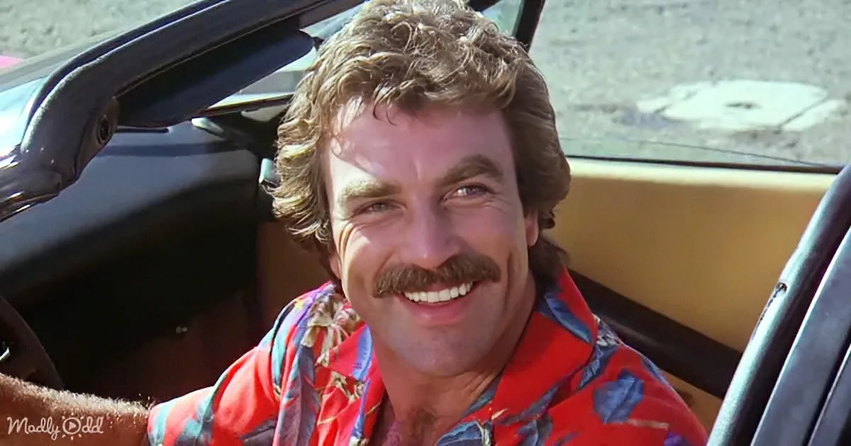 A smiling man with a mustache and curly hair sits in a convertible, wearing a colorful shirt.