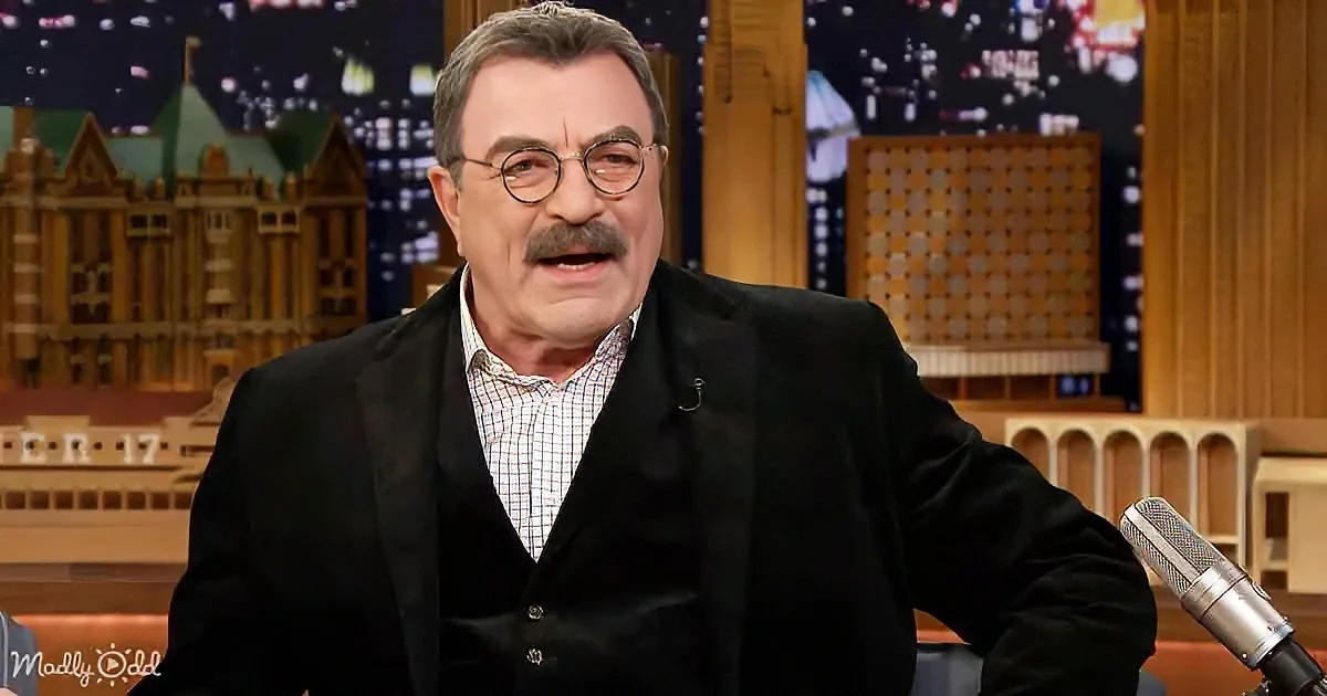 Tom Selleck, wearing glasses and a black suit, sits on a talk show set with a cityscape backdrop.