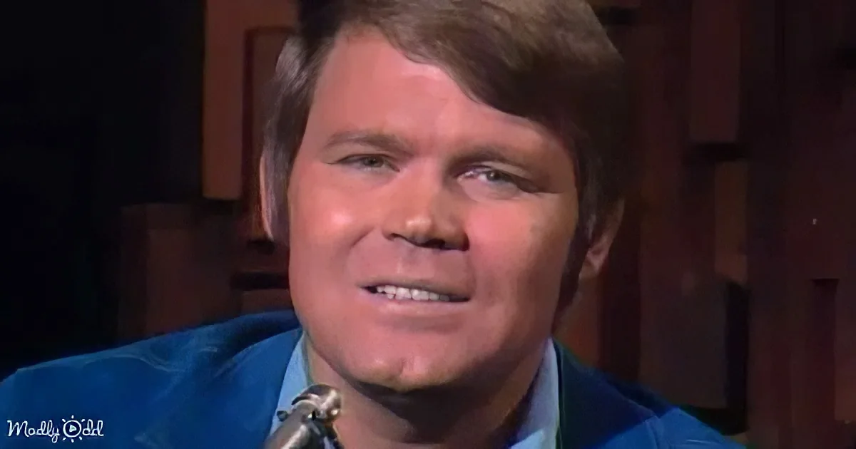 A man with short brown hair, wearing a blue suit, smiles while speaking into a microphone.