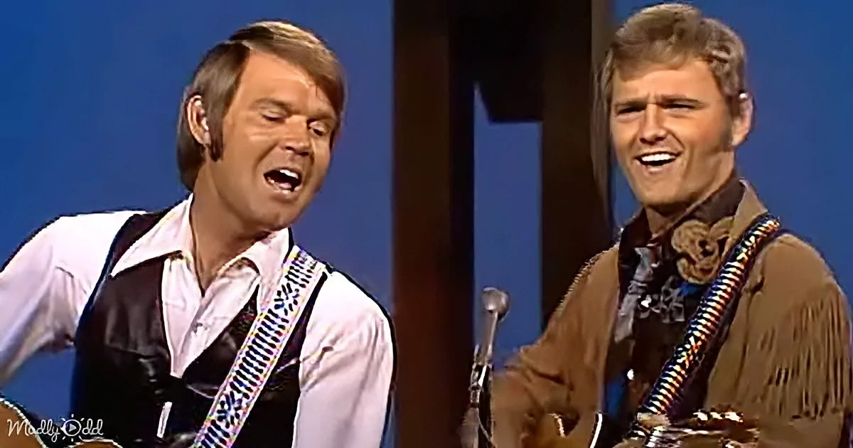 Two men in Western attire sing and play guitars on stage, one in a white shirt and black vest, the other in a brown...