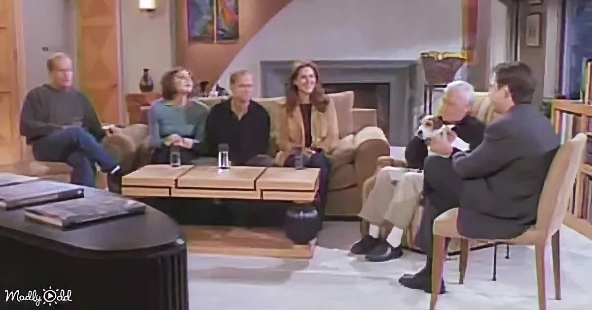A man in a suit sits in a chair, holding a dog, while others watch in a cozy living room setting.