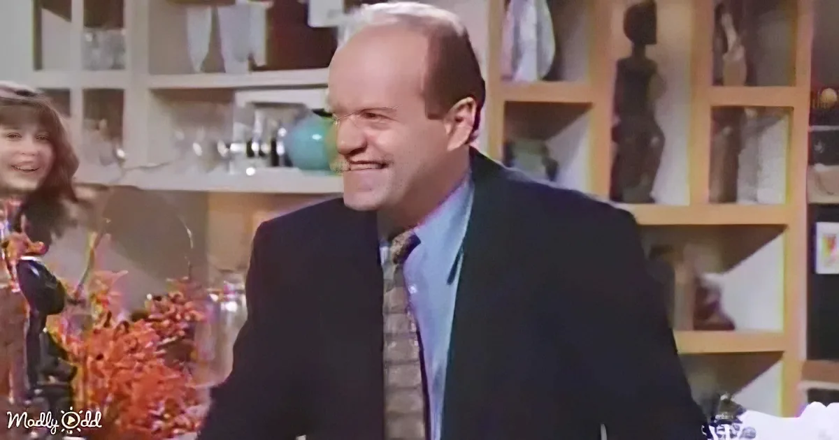 A man in a suit and tie smiles, standing in a room with shelves and decor.