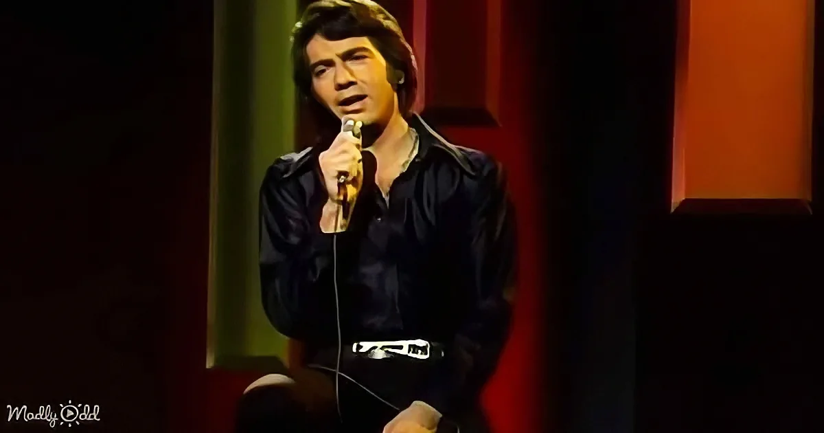 A man in a black shirt holds a microphone, standing on stage with a red curtain backdrop.