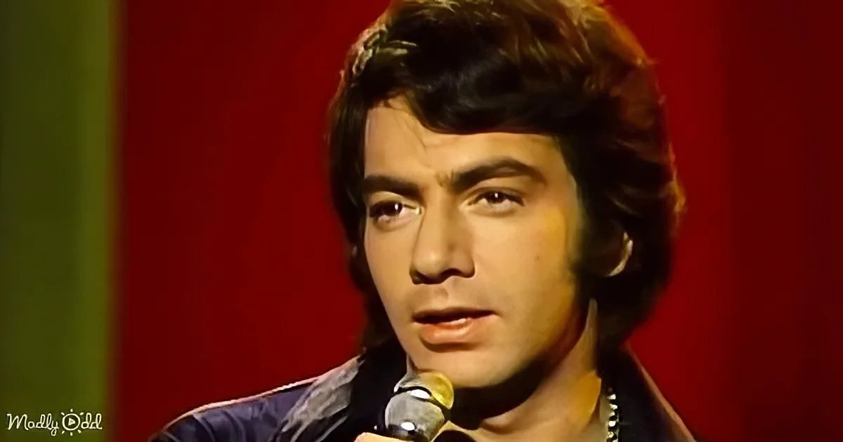 A man with dark hair and a microphone, wearing a dark jacket, against a red and green background.
