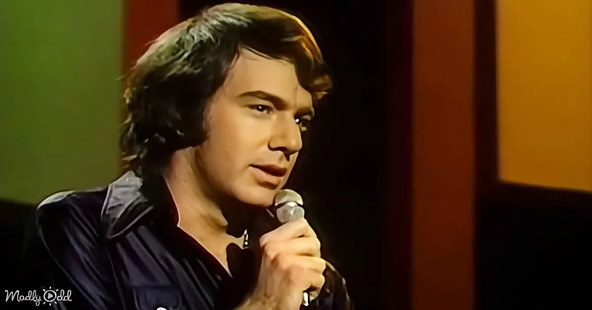 A man with dark hair, wearing a dark shirt, holds a microphone and appears to be speaking or singing.