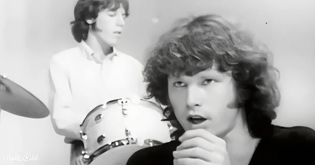 A man with curly hair, wearing a dark jacket, holds a microphone, while another person in a white shirt plays drums in...