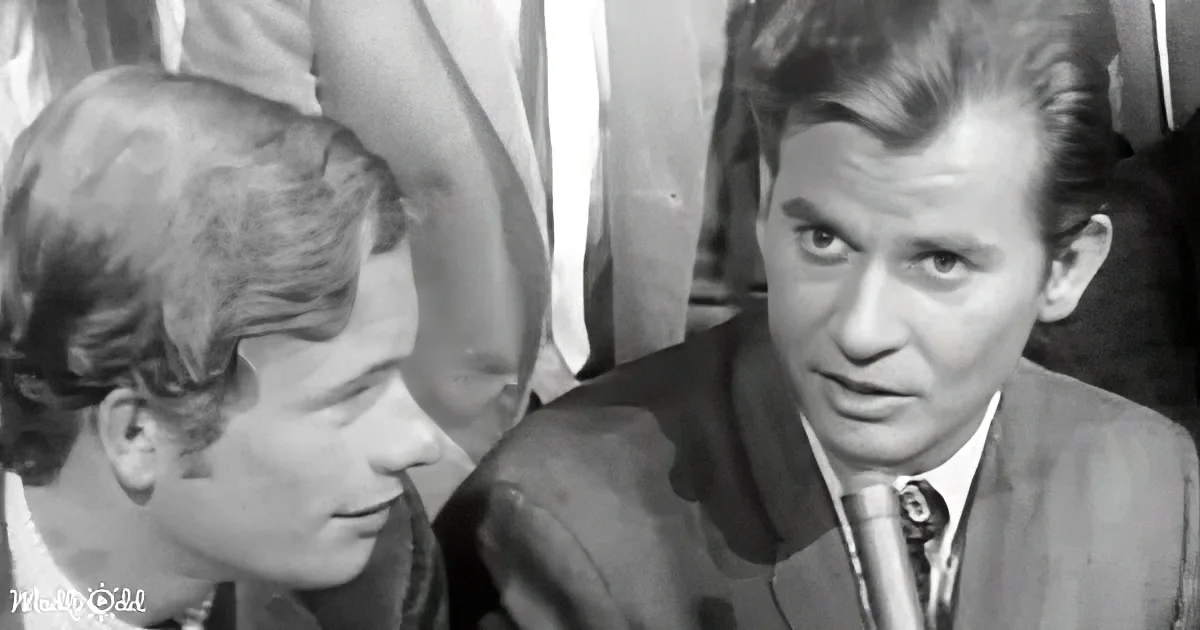 A man in a suit and tie is looking down, possibly in conversation, with another person nearby.