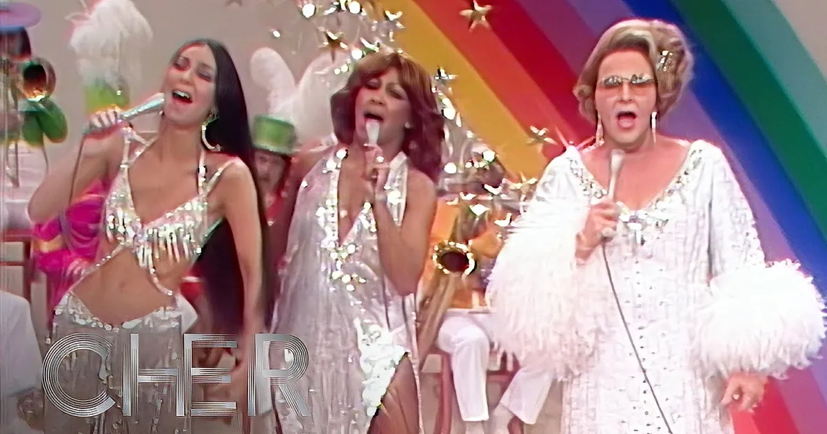 Three women in shimmering outfits perform on stage with microphones and a saxophone.