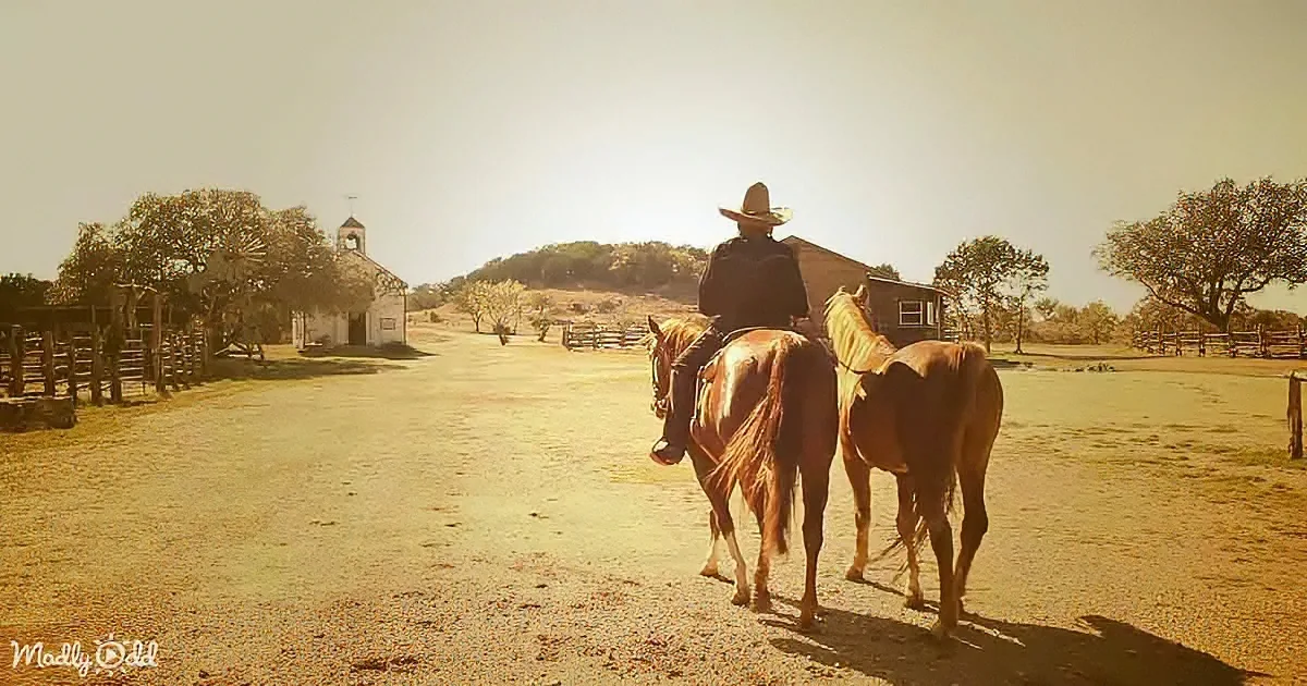 A cowboy rides two horses on a dirt road, with a church and trees in the background.