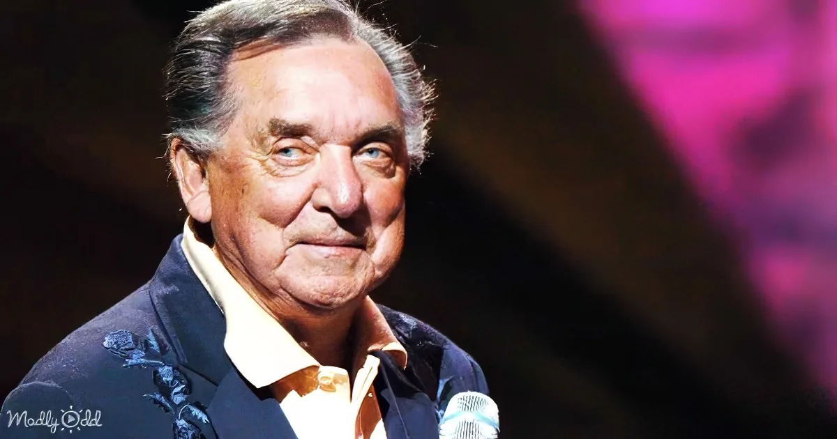 A man with gray hair, wearing a dark jacket and a yellow shirt, stands on stage with a microphone in front of him.