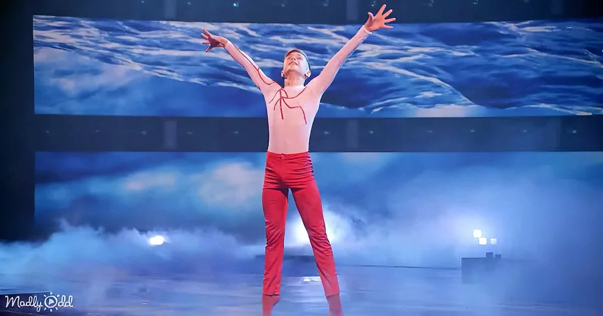 A performer in red pants and a white shirt with red lines stands with arms raised on a stage with a water-themed backdrop.