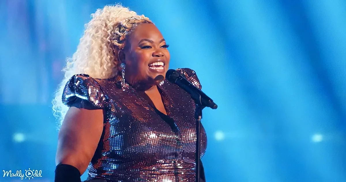 A woman with curly blonde hair in a shiny, sequined outfit sings into a microphone on stage.