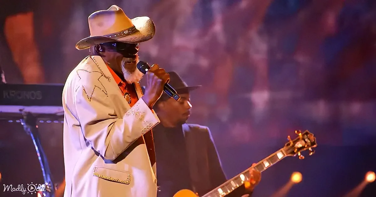 A man in a white suit and cowboy hat sings into a microphone on stage.