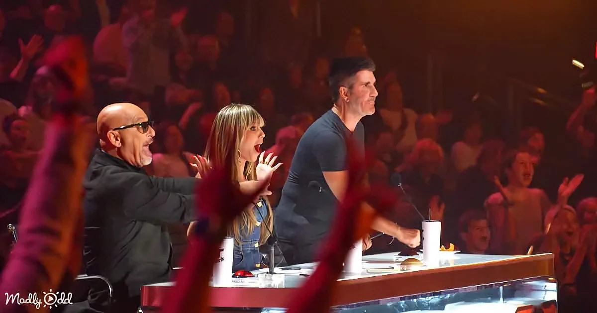 Three judges on a stage with a crowd, one holding a microphone, another gesturing, and the third clapping.