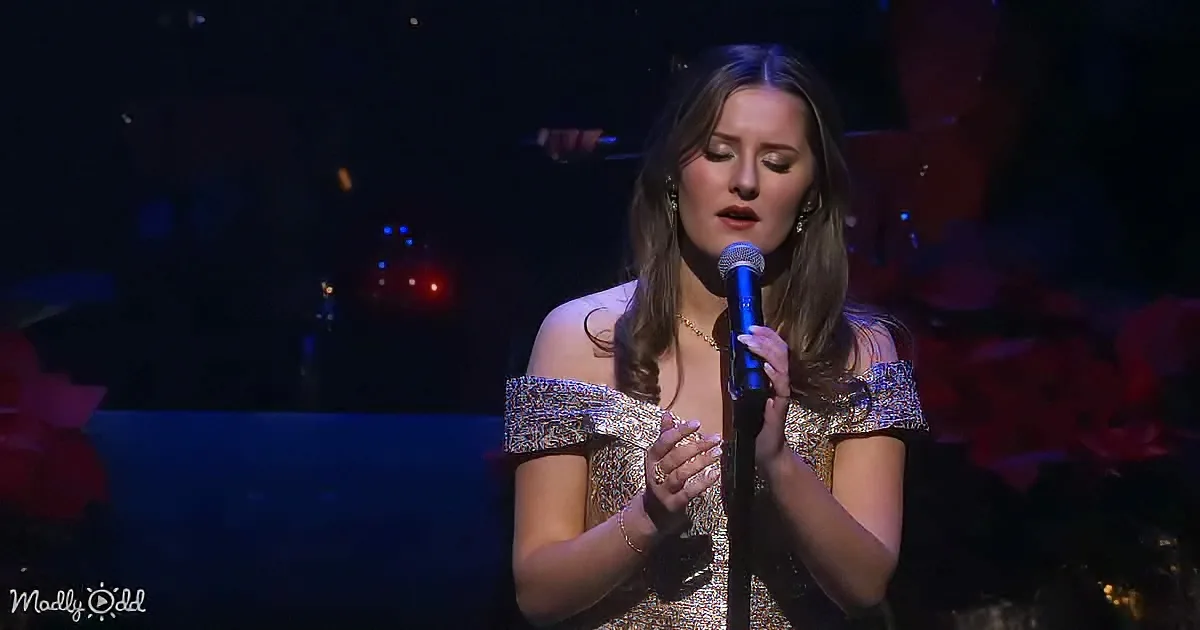 A woman in a glittery dress sings into a microphone on stage, with a dimly lit background.