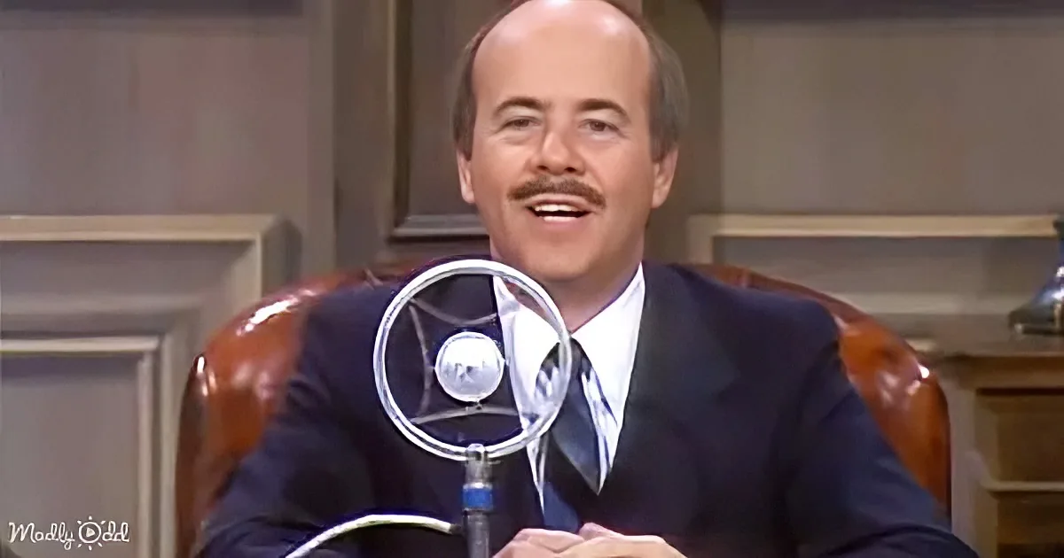 A man in a suit sits behind a microphone, smiling, in a formal setting.