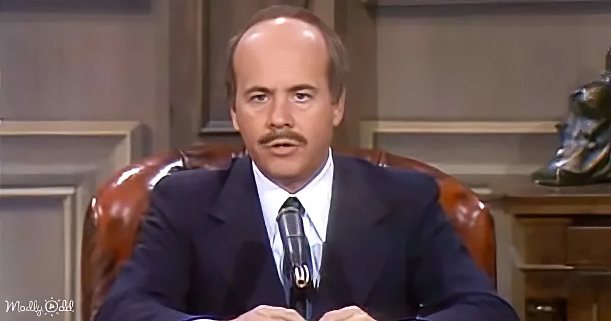 A man in a suit sits at a desk with a microphone, appearing to speak in a formal setting.