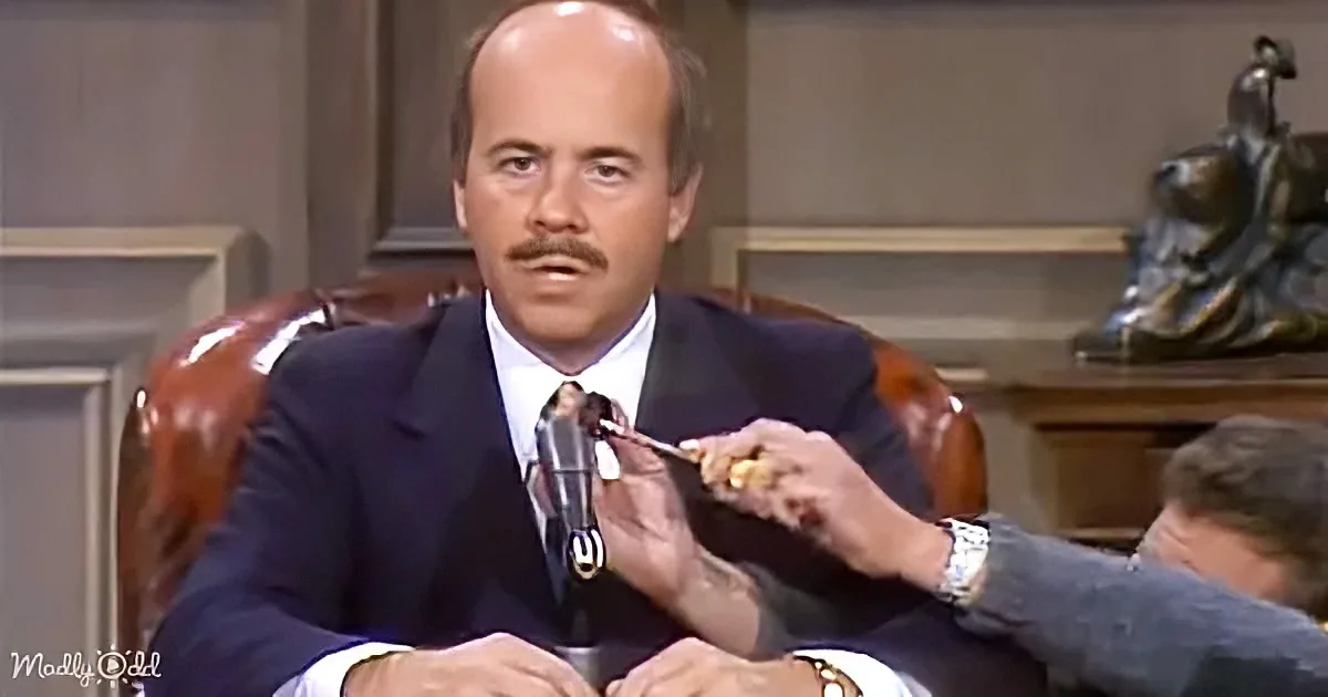 A man in a suit sits in a leather chair, holding a microphone, with another person's arm extending into the frame.