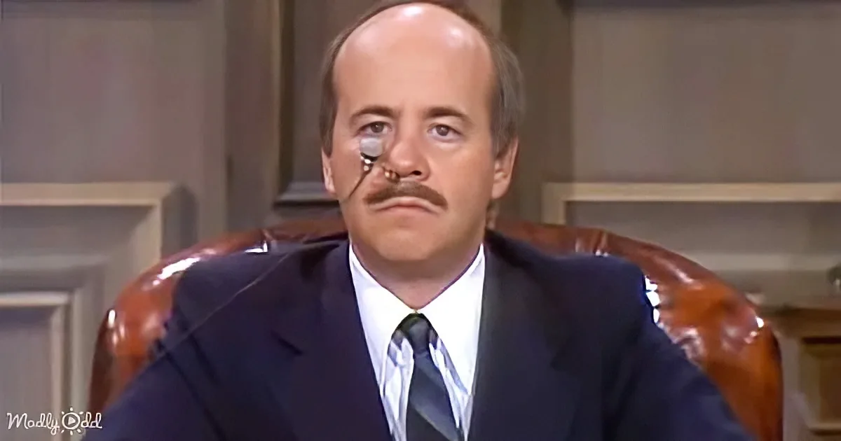 A man in a suit and tie sits in a leather chair, wearing a monocle and mustache, looking serious.