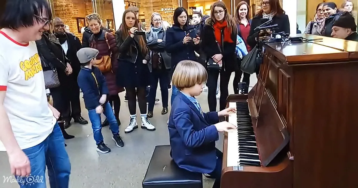 A young boy in a blue suit plays a piano while a crowd watches, including a man in a white shirt and a young boy in a...