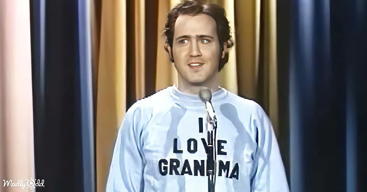 A man in a light blue shirt with "I LOVE GRANDMA" stands on stage with a microphone, looking to the side.