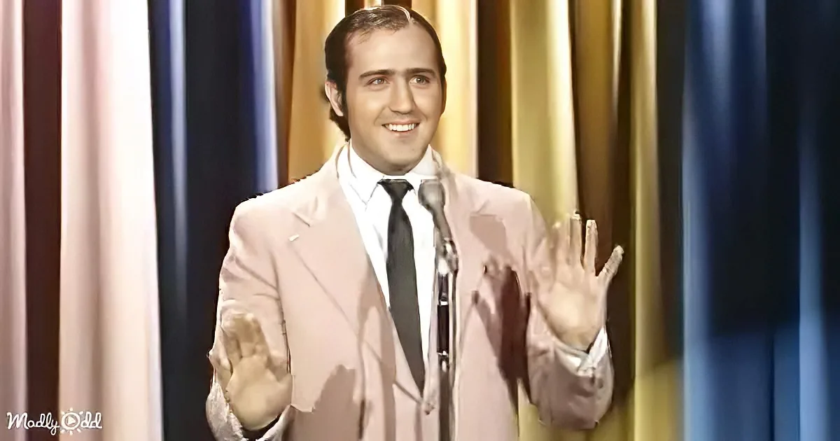 A man in a light pink suit and black tie stands on stage with his hands raised, smiling.