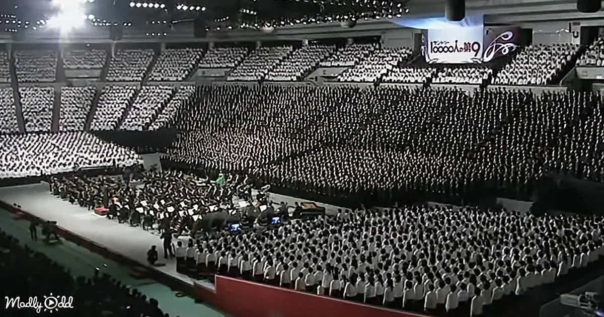 A large indoor arena filled with people in white, seated in rows, facing a stage with a conductor and orchestra.
