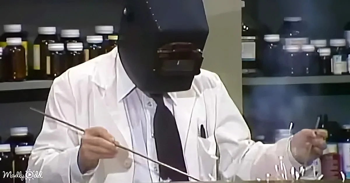 A person in a lab coat and welding mask is holding a long object and a small bottle, possibly in a lab setting.