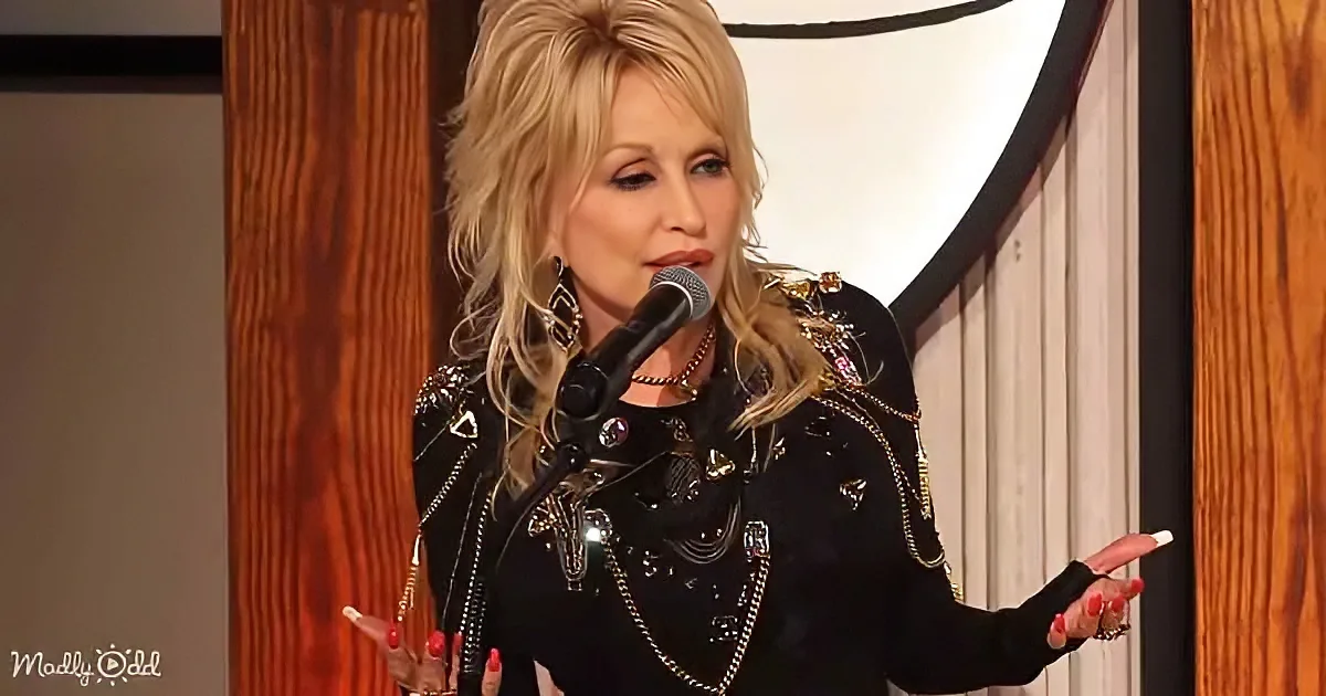 Dolly Parton, blonde, black sequined outfit, gesturing, speaking into microphone, red nails, wooden backdrop.