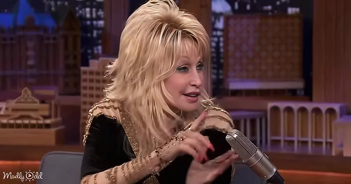 Dolly Parton, with blonde hair and a black outfit, is seated on a talk show set, gesturing with her hands.