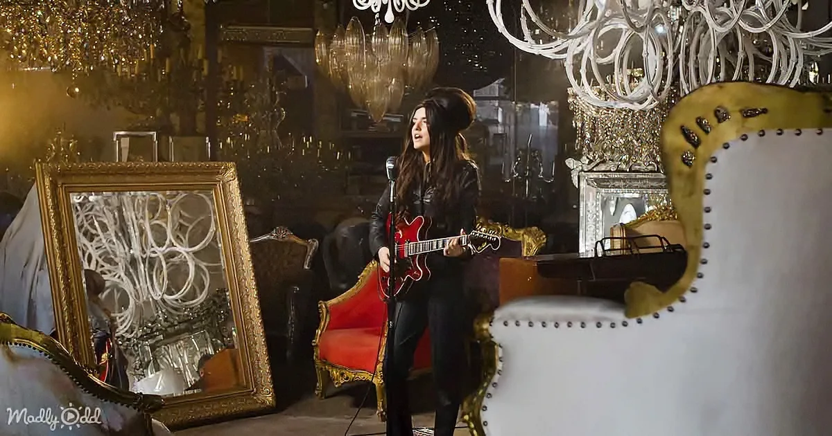A woman sings into a microphone while playing a red guitar in a luxurious, ornate room with chandeliers and vintage...