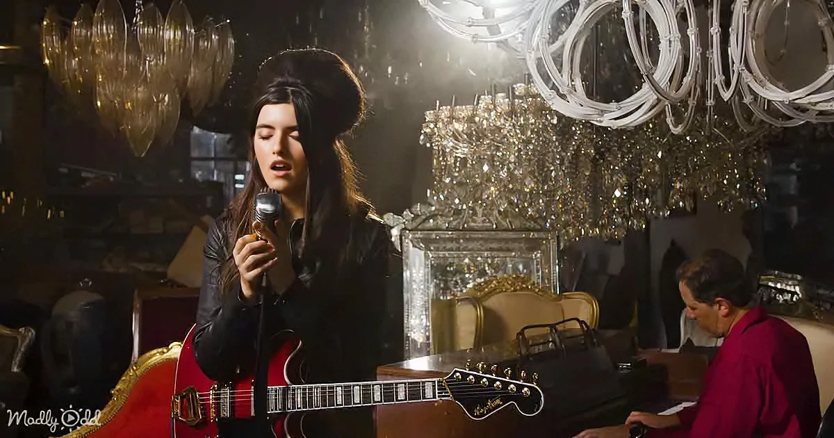 A woman sings passionately into a microphone, holding a red guitar, with a man playing piano in the background.