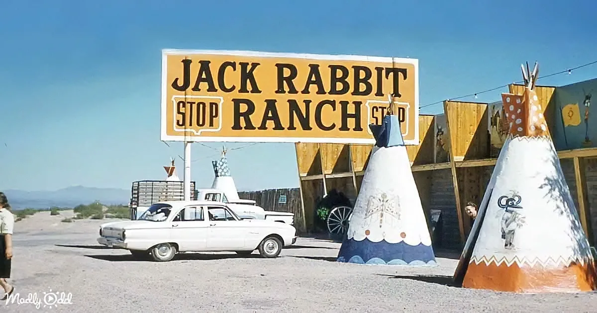 A vintage white car is parked in front of the Jack Rabbit Stop Ranch sign, with colorful teepees and a clear blue sky in...