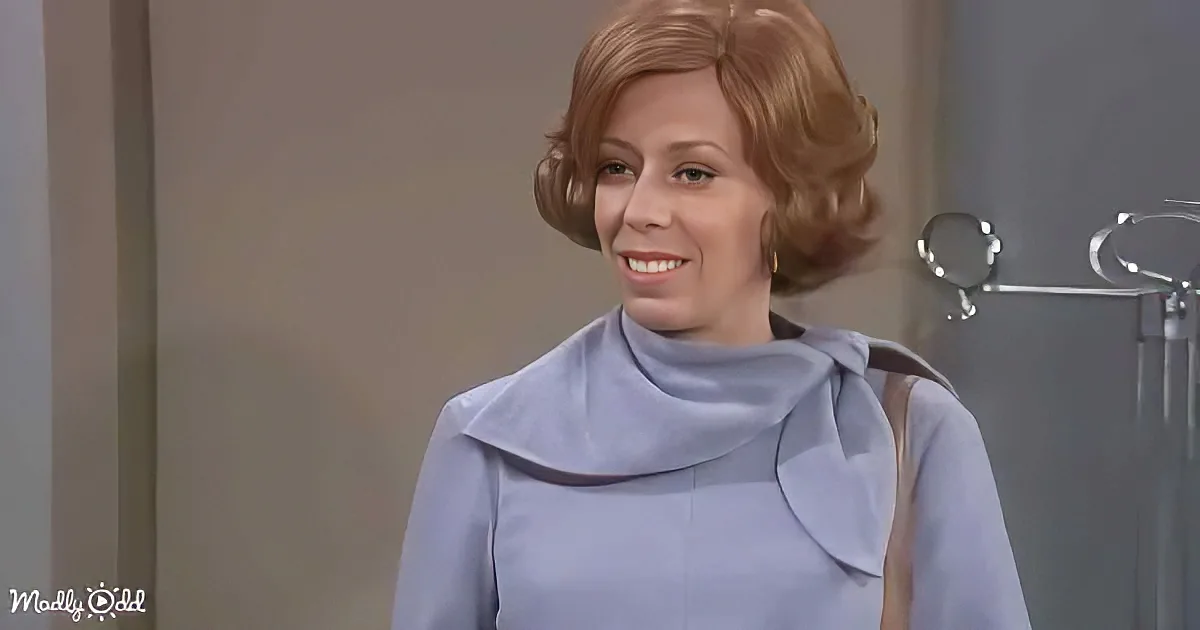 A woman with short, styled hair smiles in a light blue dress with a bow, standing in a room with a coat rack.