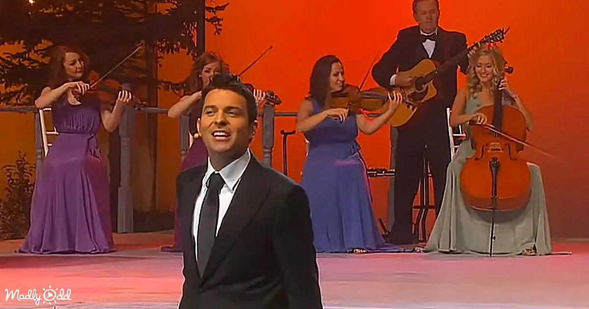 A man in a black suit sings on stage, surrounded by musicians in colorful dresses and a man in a suit playing guitar.